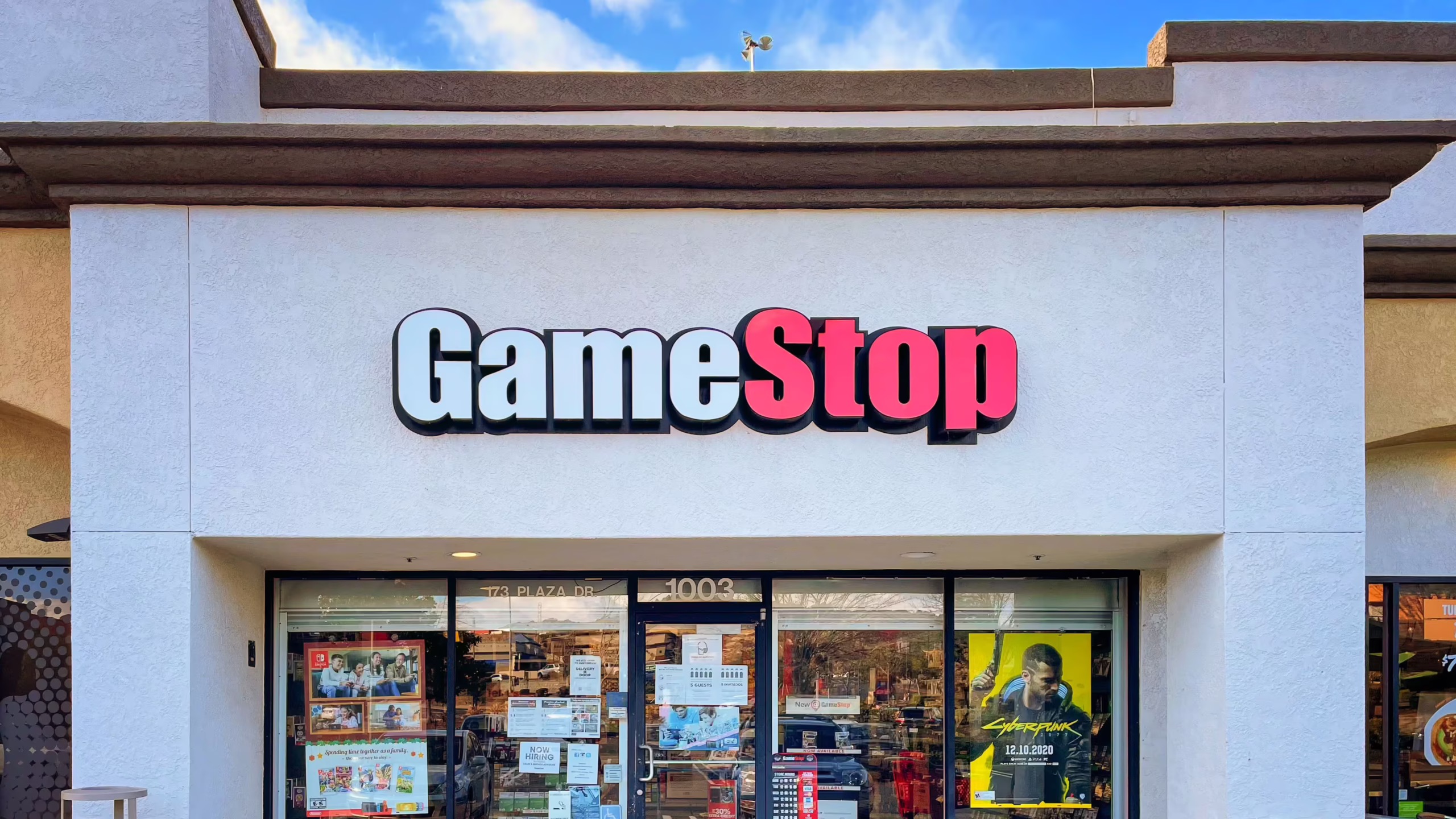 Front view of a GameStop retail store with signage, game posters in the windows, and hiring ads at the entrance.