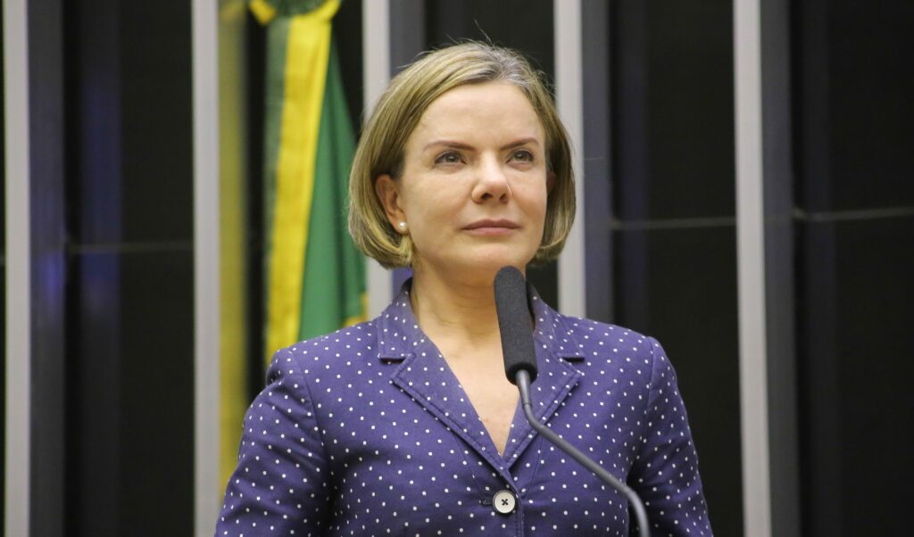 Blonde woman in a blue polka-dot blazer speaks into a microphone in the chamber, with a blurred Brazilian flag in the background.