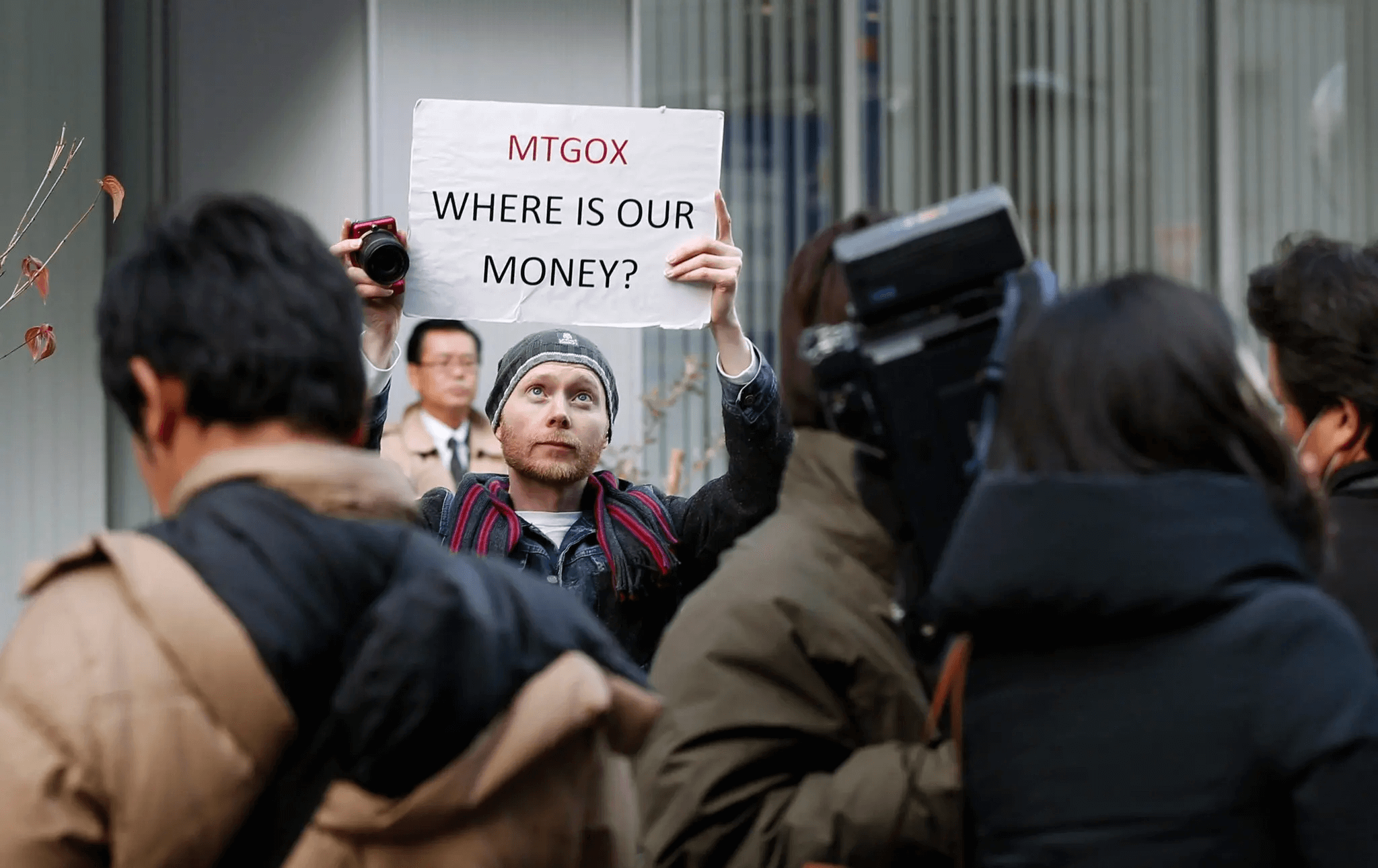 A protester holding a sign reading "MTGOX WHERE IS OUR MONEY?" during a public demonstration, surrounded by media and onlookers, referencing the Mt. Gox cryptocurrency exchange collapse