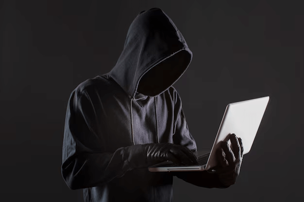 Hooded figure in dark clothing wearing gloves, using a laptop in a dimly lit setting, symbolizing cybersecurity threats or hacking