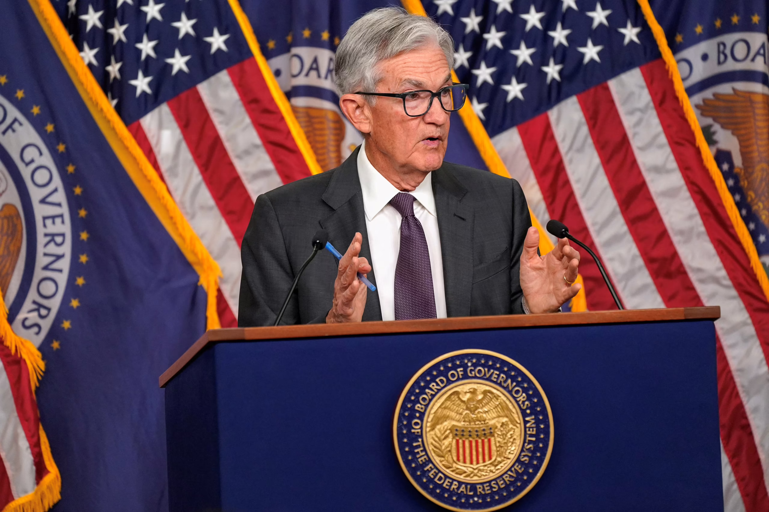 Federal Reserve Chair speaking at a press conference on interest rate policy, with U.S. flags and Fed seal in the background.
