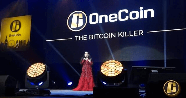 OneCoin promotional event with Ruja Ignatova on stage in a red dress, speaking under the slogan “The Bitcoin Killer” projected beside the OneCoin logo on a large screen.