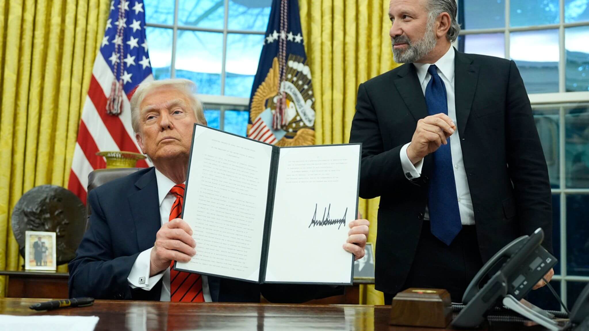 Donald Trump displays a signed executive order in the Oval Office, with US flags in the background, symbolizing official presidential action.
