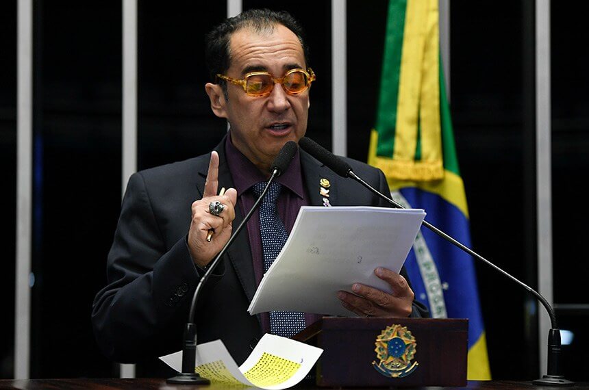 Homem de terno faz discurso em púlpito do Senado Federal do Brasil, com documentos em mãos e a bandeira nacional ao fundo.