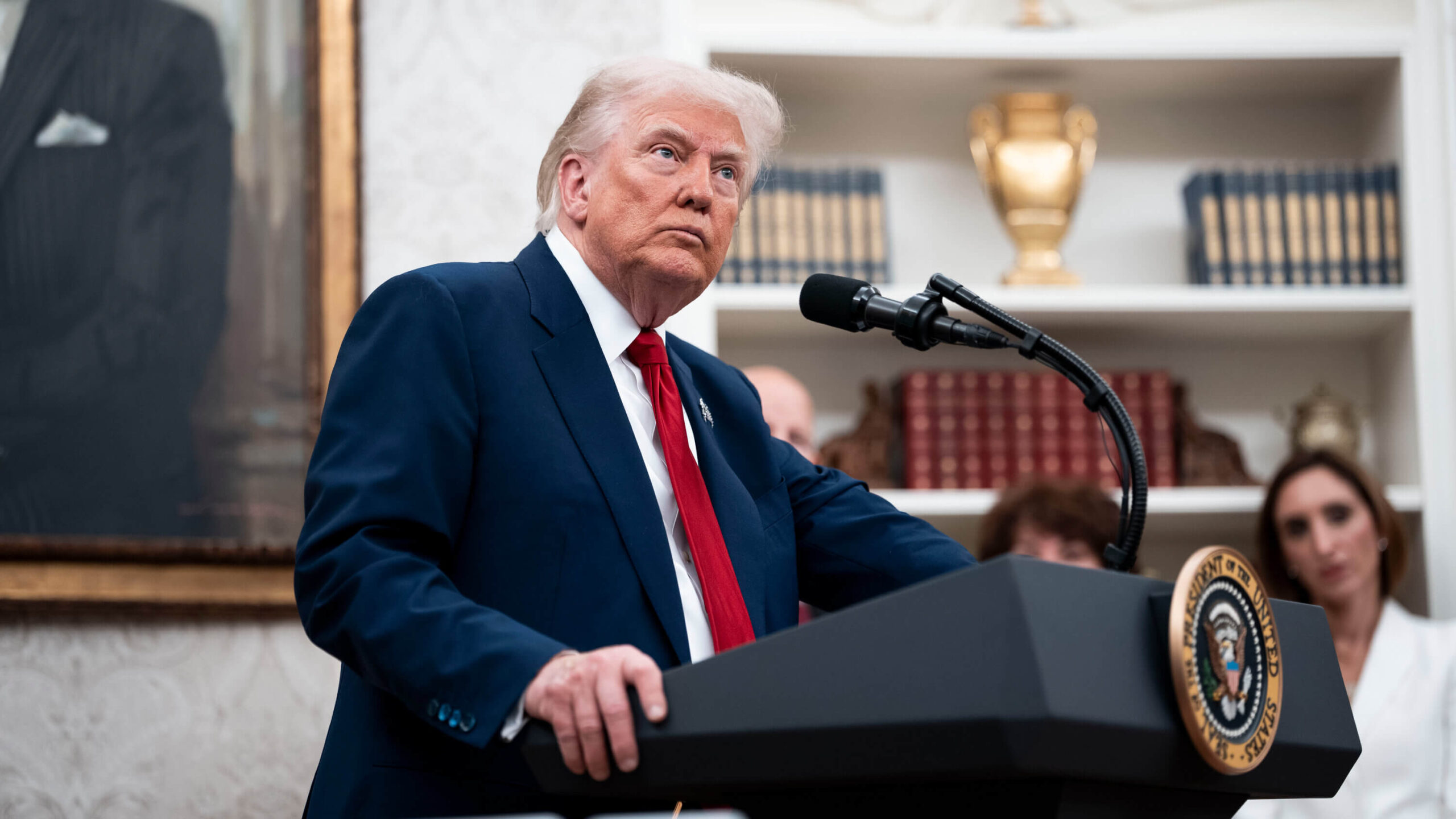 Trump in a blue suit and red tie delivers an official speech at the US presidential lectern, with books and listeners in the background.