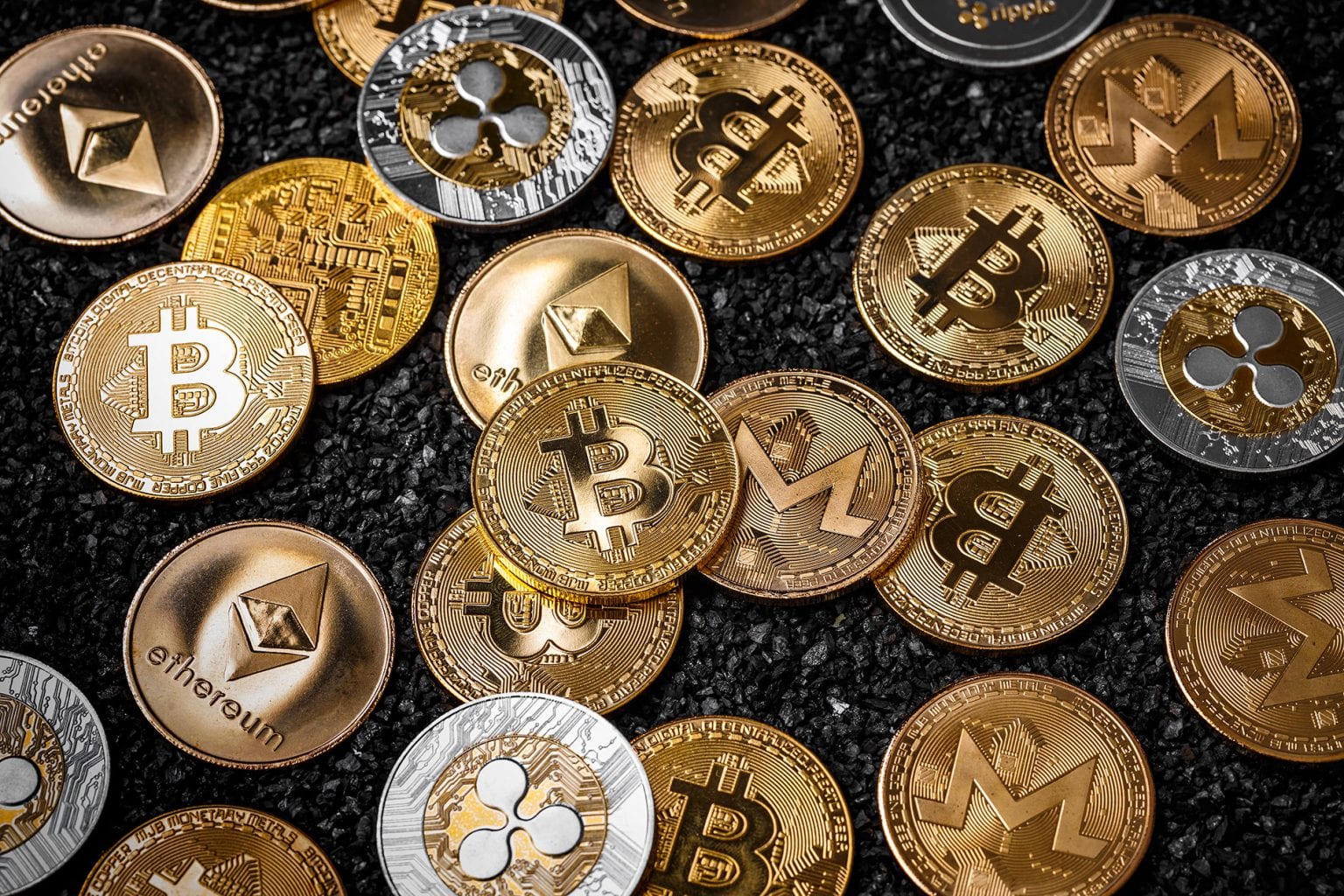 Diversas moedas físicas de criptomoedas como Bitcoin, Ethereum, Monero e Ripple sobre fundo escuro, simbolizando o mercado cripto.